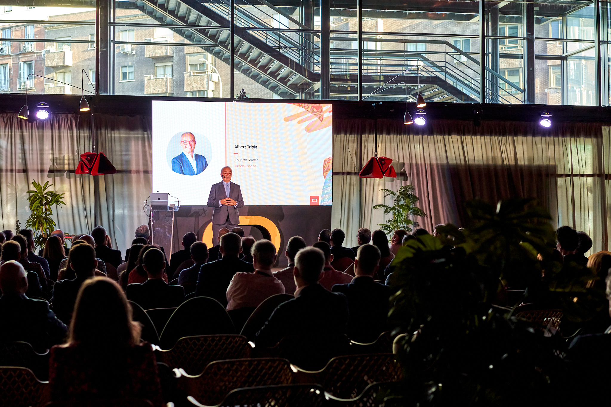 Fotógrafo de eventos corporativos en Madrid cubriendo una jornada. Muestra a un ponente en el escenario, con su nombre (Albert Triola) visible en la gran pantalla. La sala es moderna, acristalada, con luz natural y detalles industriales. Ideal para reportaje de conferencias y eventos para empresas.