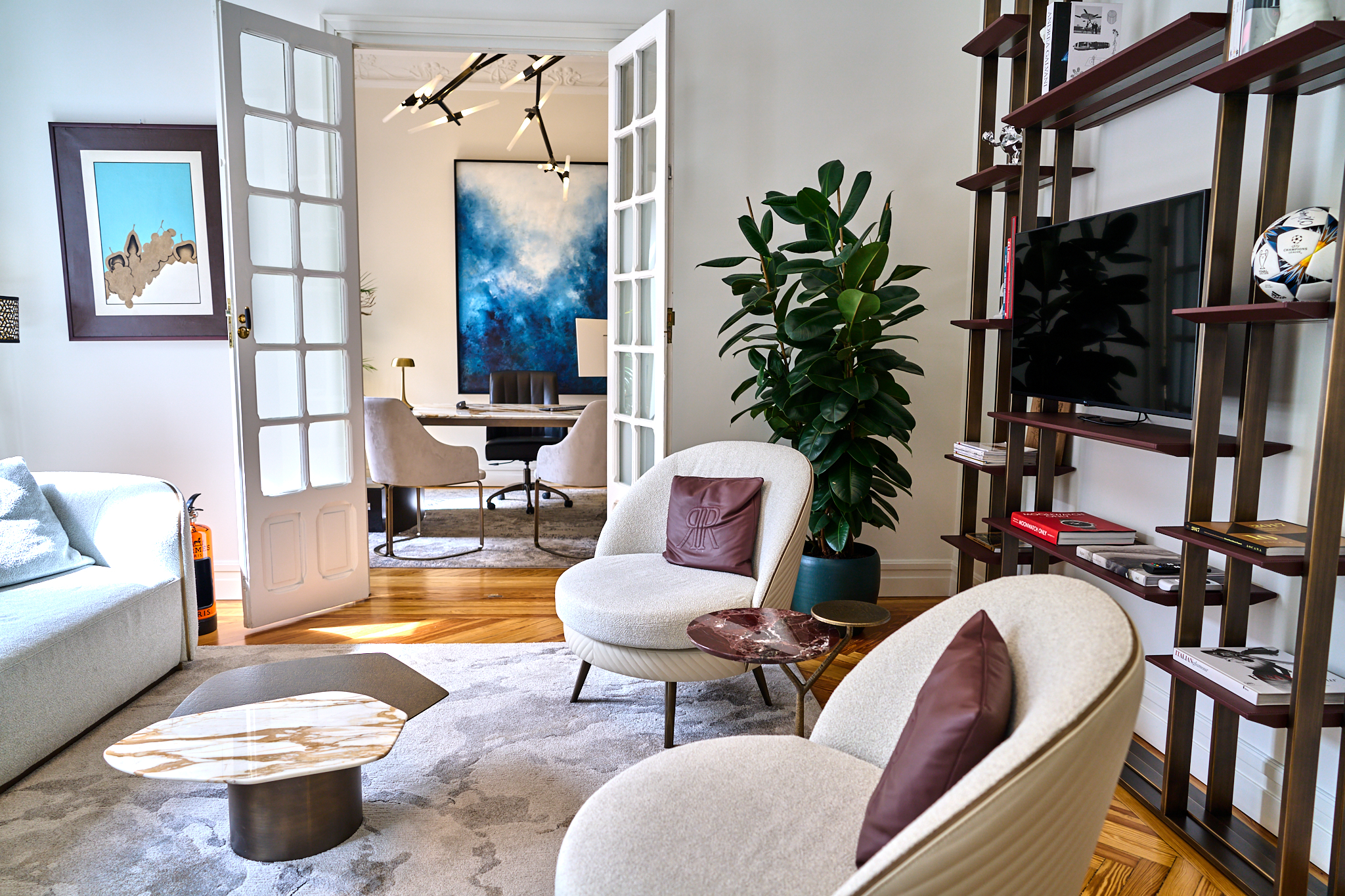 Fotografía corporativa de reportaje de instalaciones y facilities de una oficina de alto standing en Madrid (Scaena Capital). Muestra un salón con muebles de diseño, grandes puertas francesas y un despacho moderno al fondo con arte abstracto azul.