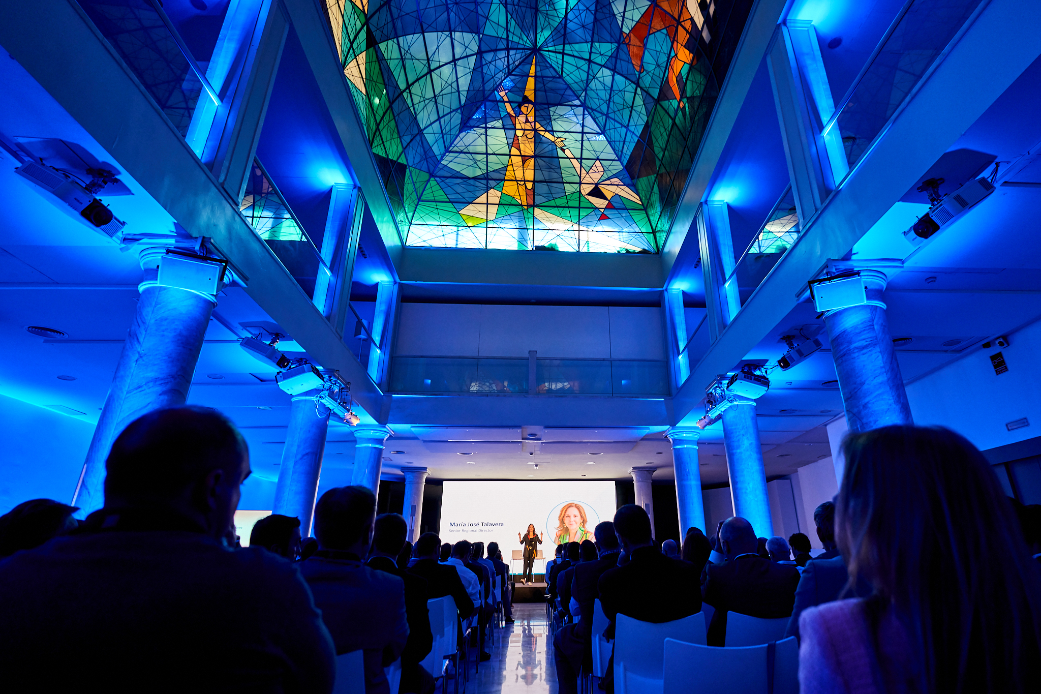 Fotografía de congreso o jornada en Madrid. Fotógrafo profesional en Madrid cubriendo un evento en un salón de lujo con vidrieras artísticas, destacando la asistencia y la iluminación azul. Ideal para fotografo para empresas.