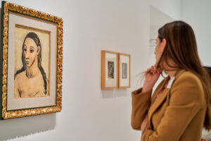 Fotógrafo de eventos en Madrid cubriendo un evento cultural o visita de empresa a un museo o galería. Muestra a una mujer mirando un cuadro enmarcado con un marco dorado sobre una pared blanca. Ideal para reportaje de marca en un contexto artístico