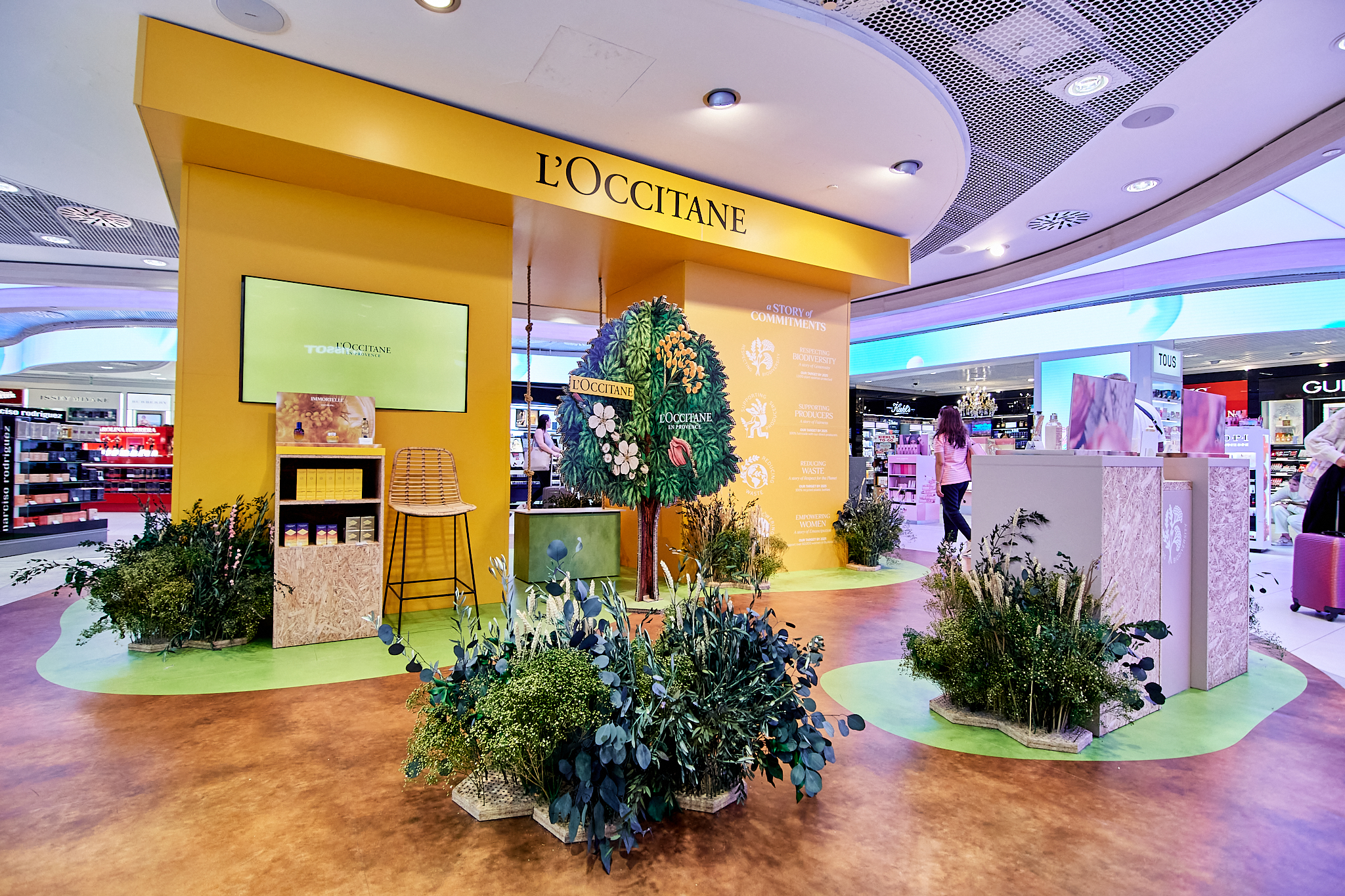 Fotografía de reportaje de stand promocional de L'Occitane en el aeropuerto de Madrid (Duty-Free). Muestra un diseño temático con elementos naturales, plantas y tonos amarillos. Ideal para cobertura de ferias, pop-up stores y activaciones de marca.